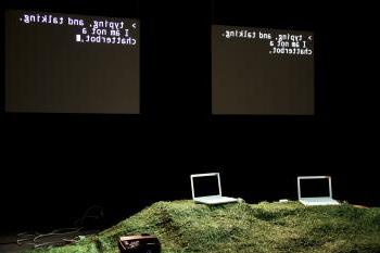 在黑暗的舞台上，两台笔记本电脑放在长满草的小山上. 在他们身后, 有两个黑屏，上面写着“打字和说话”, 我不是一个聊天机器人”.